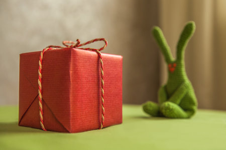 Red gift box with decorative rope bow in foreground with blurred green crochet bunny sitting in background on tableの写真素材