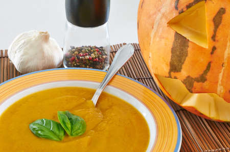 A bowl of pumpkin soup with a couple of basil leaves, pepper, garlic and a Halloween pumpkinの写真素材