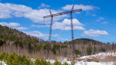 The support of an overhead power line among mountains and forests, under a blue sky with clouds.