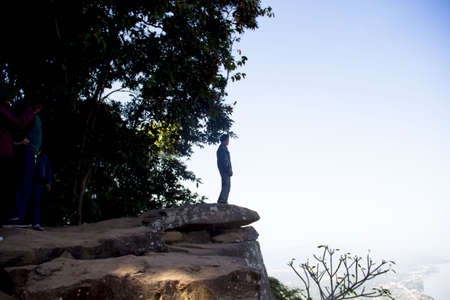 Young man in black sportswear is sitting on cliff's edge and looking to misty valley bellow