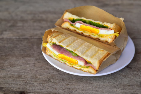 Sandwich cut in half. Two pieces of sandwich with ham, egg, lettuce, tomatoes and sauce in a white plate on old wood table. Top view, copy spaceの写真素材