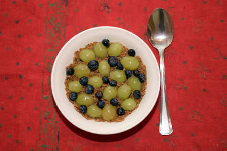 A bowl of cereal with sliced grapes and whole blueberries on top displayed on a Christmastime table cloth.の写真素材