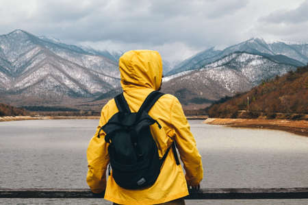Backpacker in yellow raincoat with backpack standing on the bank of mountain lake and looking at snowy mountains.の写真素材