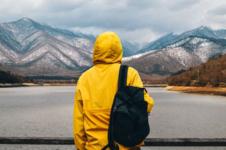 A man in a yellow jacket with a backpack stands on the shore of a mountain lake and looks at the mountainsの写真素材