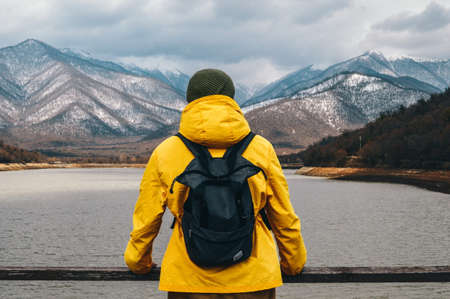 A man in a yellow raincoat with a backpack stands on the bank of a mountain river and looks at the snowy mountains.の写真素材