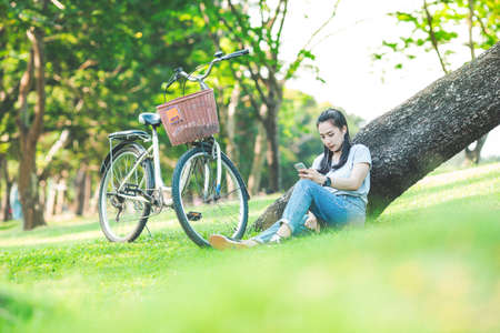 woman sitting in a park leaning on a tree and listening to music on headphones with a bicycle.Image with Grain.の写真素材