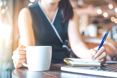 The hand of a business woman taking notes in a notebook in the office.And another hand holding a cup of coffee.の写真素材
