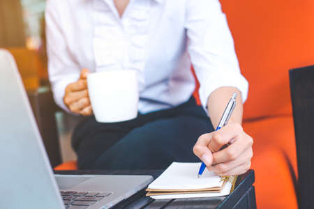 The hands of a business woman are writing with a pen on a notepad and holding a cup of coffee.