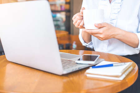 Business woman hand holding a cup of coffee.On her desk is a notebook computer,cellphone Pen and notepadの写真素材