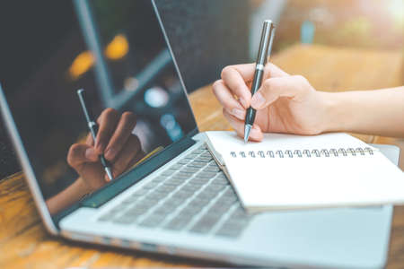 A woman's hand working on a notebook computer and writing on a notepad with a pen in the office.
