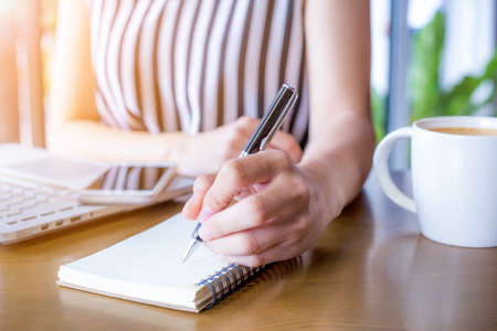 Business woman hand working at a computer and writing on a noteped with a pen in the office.On the wooden desk there is a cup of coffee and a mobile phone.の写真素材