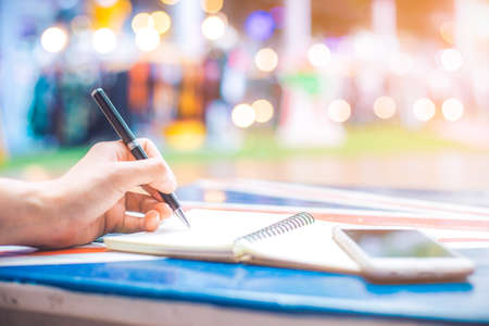 Woman's hand writing on a notebook with a pen on a wooden desk.On the table are mobile phones.Background blur backlightの写真素材