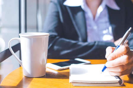 business woman hand  writing on notepad with a pen in office.On her wooden desk there are cell phones and coffee cups.の写真素材
