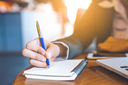 business woman hand  writing on notepad with a pen in office.On the wooden table there is a laptop computer and a mobile phone.の写真素材