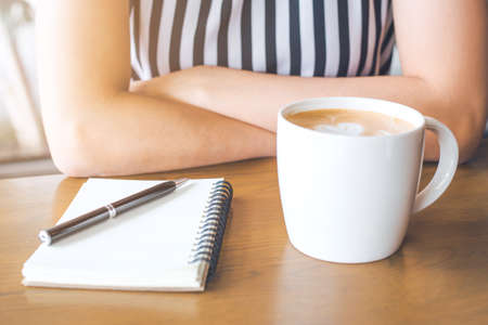 A woman's hand sitting on a coffee table on a table with a cup of coffee, notepad and pen.の写真素材