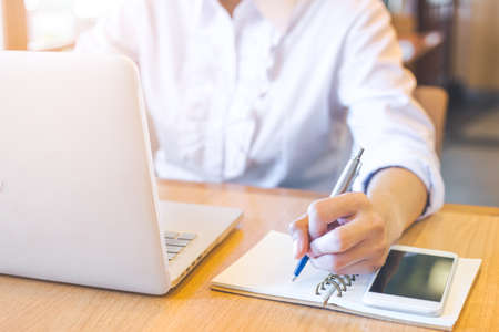 A business woman hand working on a laptop computer and writing on notepad with a pen on her office...