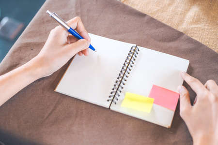 Hand is writing on a blank notepad with a pen, on a brown tablecloth.