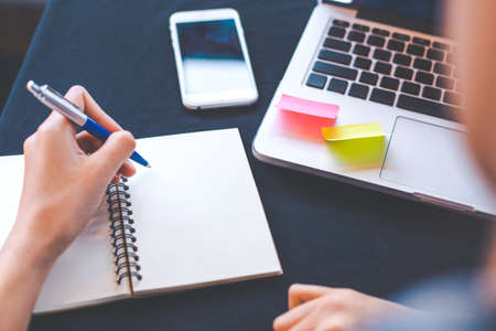 Woman hand works in a laptop computer and is writing on notepad with a pen in the office.On her desk there is a cell phone.の写真素材