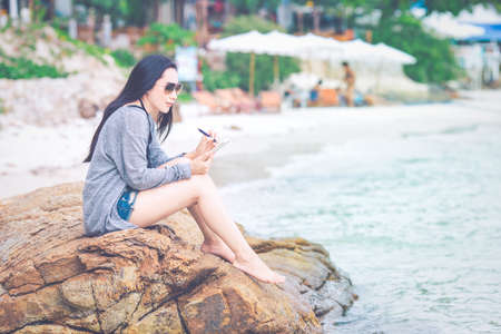 Woman writing on notepad with a pen at the beach.She sits at the rocks and wears sunglasses.の写真素材