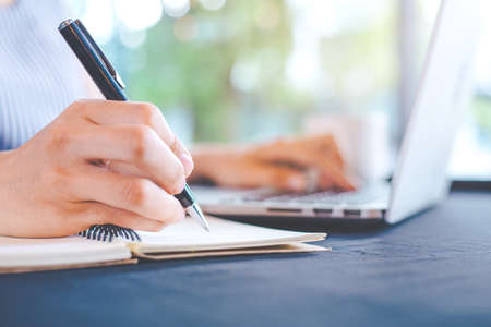 Woman's hand writing on notepad with a pen and works in a laptop computer in the office.の写真素材