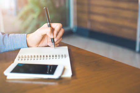 Female hand is writing on notepad with pen in office.On the wood table there are cell phones.