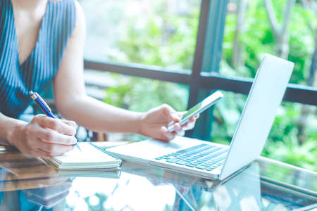 Business woman hand is writing on a notepad with a pen and using a mobile phone.On the table is a laptop computer.の写真素材