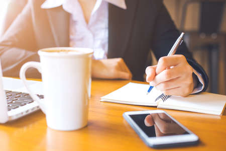 Business woman hand writing on notepad with a pen in office.On her wooden desk there are cell phones.の写真素材
