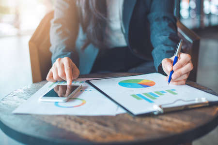 Business woman hand uses a phone to work on charts and graphs that show results.She uses the pen to point to the graph.の写真素材