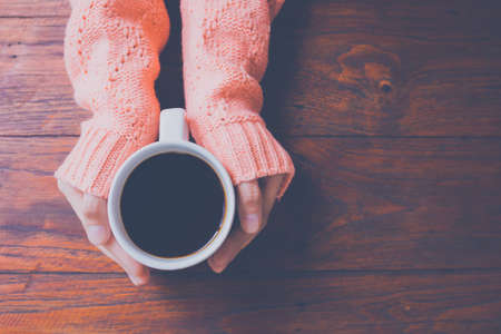 Woman hand in warm sweater holding a cup of coffee on a wooden table background, top viewの写真素材