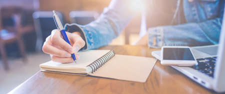 A woman's hand is writing in empty spiral notepad with a pen.On the table are the phones that are on the laptop computer.Web banner.の写真素材