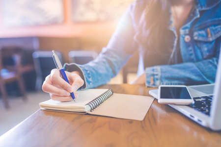 A woman's hand is writing in empty spiral notepad with a pen.On the table are the phones that are on the laptop computer.の写真素材