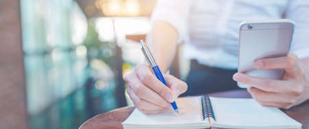 Woman hand writing on a notepad with a pen and using a mobile phone in the office.Web banner.の写真素材