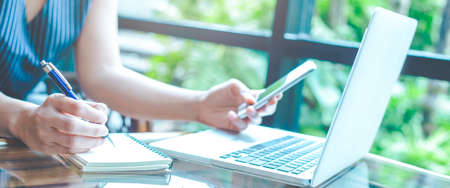 Business woman hand is writing on a notepad with a pen and using a mobile phone.On the table is a...