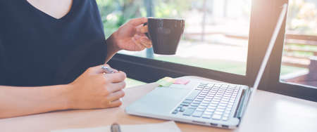 Woman hand is using a laptop computer in office.In her hand holding a cup of black coffee.Web banner.の写真素材