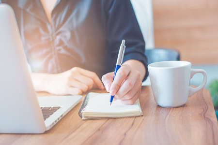 Business woman is writing on a notebook with a pen and using a laptop to work in the office.On a wooden table with a cup of coffeeの写真素材