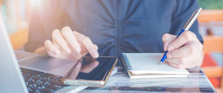 Woman is writing on a notebook with a pen and she is using a mobile phone and computer laptop in the office.の写真素材