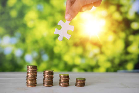 Man's hand holding  piece of puzzle with Stacks of gold coins on wooden table with green bokeh background.Money solution concept.の写真素材