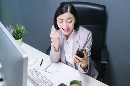 Happy chinese female worker at her office desk working on her computer. and smartphone.Excited face expression.の写真素材