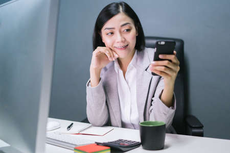 Happy chinese female worker at her office desk using her smartphone and smiling.の写真素材