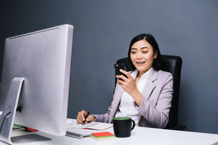 Happy chinese female worker at her office desk using her smart phone and smiling.の写真素材