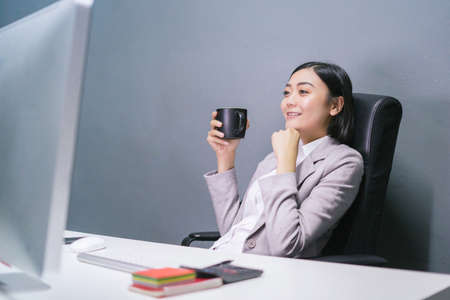 Happy asian female worker at having coffee at her office desk and smiling.の写真素材