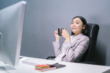 Happy asian female worker at having coffee at her office desk and smiling.の写真素材