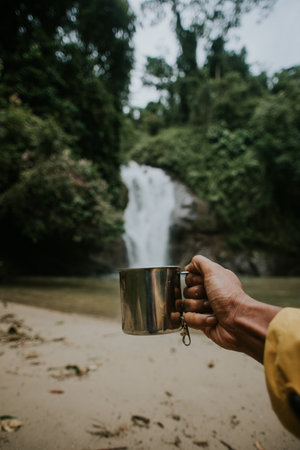 Close up shot of a man's hand holding coffee mug with waterfall in the background.Chilling during camping and hiking.の写真素材