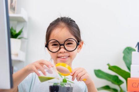 Cute little asian girl holding a magnifying glass and watering a plant in the room