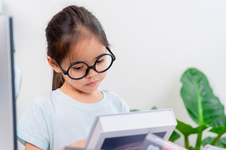 Cute little asian girl with eyeglasses reading book at homeの写真素材