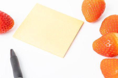 A yellow notepad with a black pen sits on a white surface. On the surface, there are several strawberries, some of which have been cut in halfの写真素材
