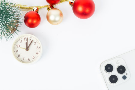 A white iPhone sits on a table next to a clock and a bunch of red and gold Christmas ornamentsの写真素材