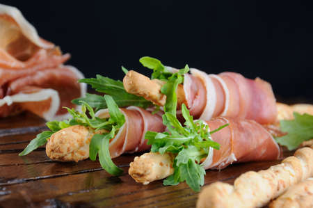 Grissini -   bread sticks with Parmesan, wrapped with a piece of prosciutto and arugula. Italian dish with antipast on a wooden table.の写真素材