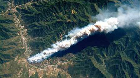 Large wildfire raging through forest near populated areas, generating thick plumes of smoke,...