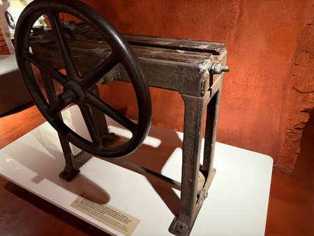 Vintage cast iron industrial press with a large handwheel standing on a white pedestal, showcasing historical manufacturing technology in an exhibit against a rustic wallの写真素材